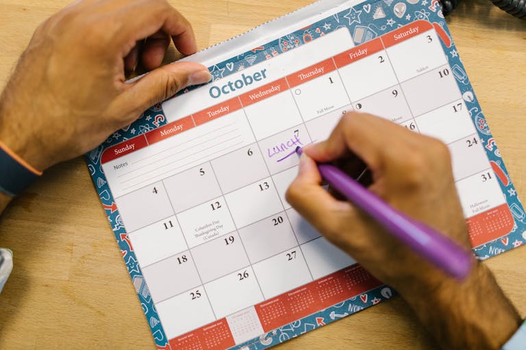 Close-up of a person writing a lunch reminder on an October calendar with a purple pen.